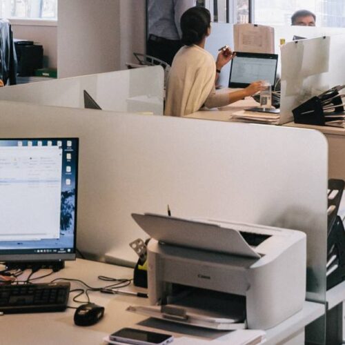 Modern open-plan office with several people working at desks with computers. A printer, keyboard, and monitor with emails open are visible in the foreground. Practice printer maintenance to extend printer life amid busy workdays and scattered supplies.