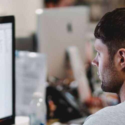 A person with short dark hair sits at a desk, focused on a computer screen displaying a design or website layout in an office setting, where printer refurbishing takes place amid blurred background elements.