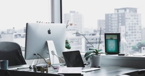 A modern office desk with an iMac, a laptop, a new printer, a potted orchid, a cup, and framed artwork sits by large windows overlooking a cityscape with tall buildings.