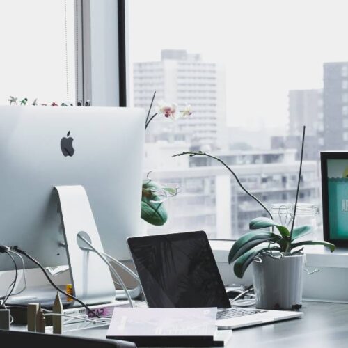 A modern office desk with an iMac, a laptop, a new printer, a potted orchid, a cup, and framed artwork sits by large windows overlooking a cityscape with tall buildings.
