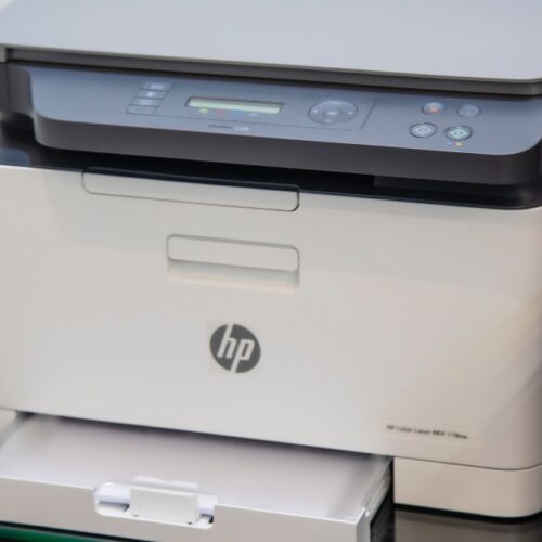 A modern HP all-in-one printer, ideal for comparing inkjet vs laserjet options, with a control panel and open paper tray, sits on a glass surface near a wall.