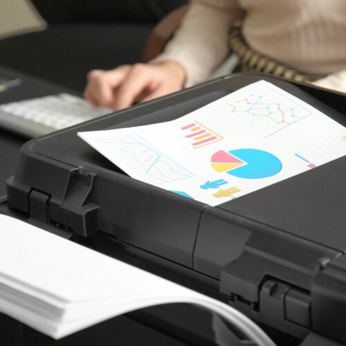 A color printer outputs a page with graphs and charts; in the background, a person types on a keyboard at a desk, showcasing the benefits of HP Printers for productive and vibrant office workflows.