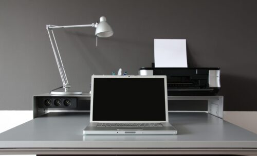 A minimalist desk setup with a closed laptop, silver desk lamp, pens in a holder, and a home office printer ready for use—a perfect example of reasons to upgrade your printer for efficiency and style against a sleek gray wall.