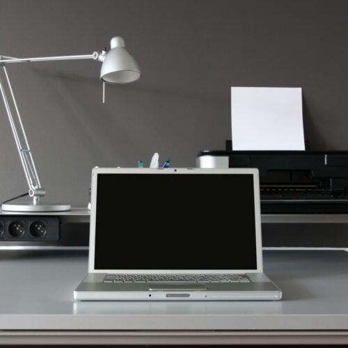 A minimalist desk setup with a closed laptop, silver desk lamp, pens in a holder, and a home office printer ready for use—a perfect example of reasons to upgrade your printer for efficiency and style against a sleek gray wall.