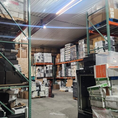 A cluttered warehouse with shelves stacked high with boxes and refurbished printers, along with various office equipment. The floor is partially obstructed by more printers and assorted items. Overhead lighting casts a dim glow, highlighting the packed shelves and scattered goods.