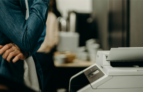 A person in a denim jacket stands with crossed arms next to a copy machine, looking like theyre pondering the top 10 deals on printers. The scene is indoors, featuring a blurred background with another individual and some office supplies.