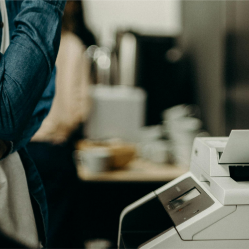 A person in a denim jacket stands with crossed arms next to a copy machine, looking like theyre pondering the top 10 deals on printers. The scene is indoors, featuring a blurred background with another individual and some office supplies.