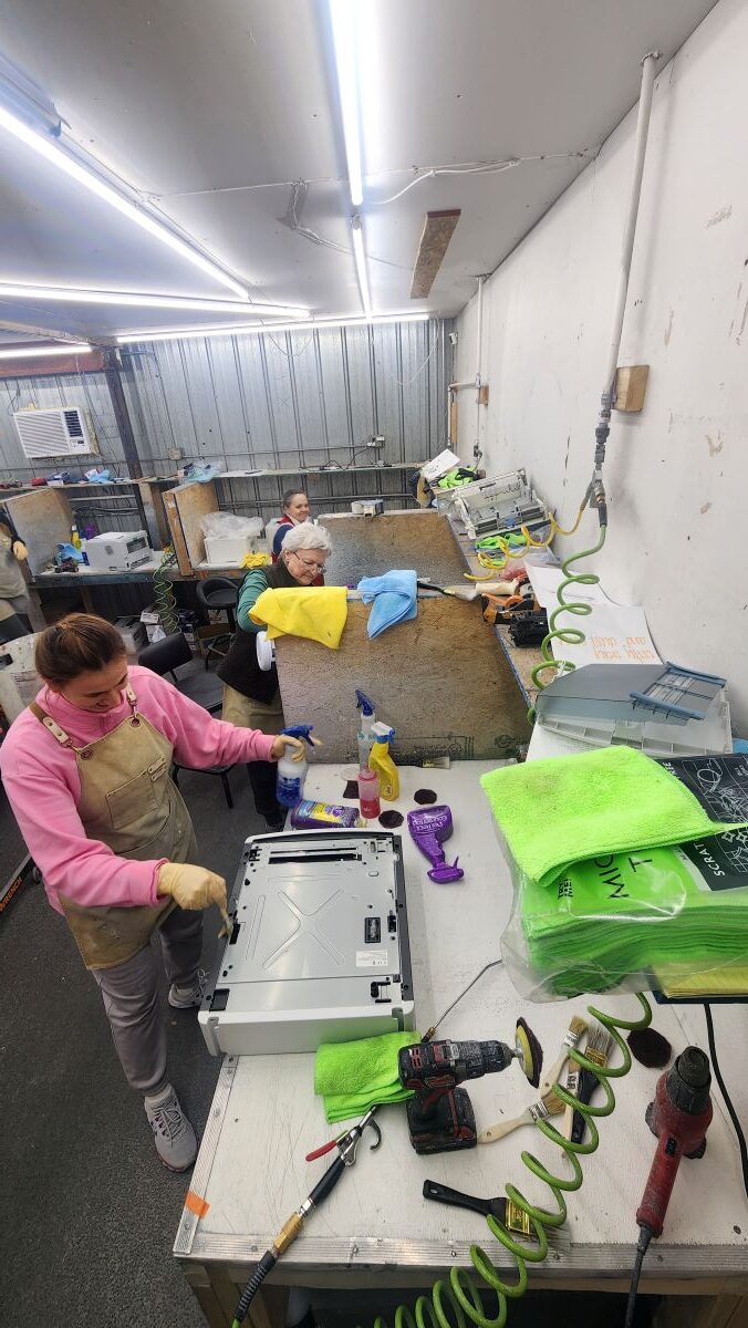 In a fluorescent-lit workshop, people are focused on refurbishing printers, diligently working on machinery and cleaning parts. Supplies like cloths, cleaning sprays, and tools are neatly arranged on tables as they meticulously restore each unit to working order.