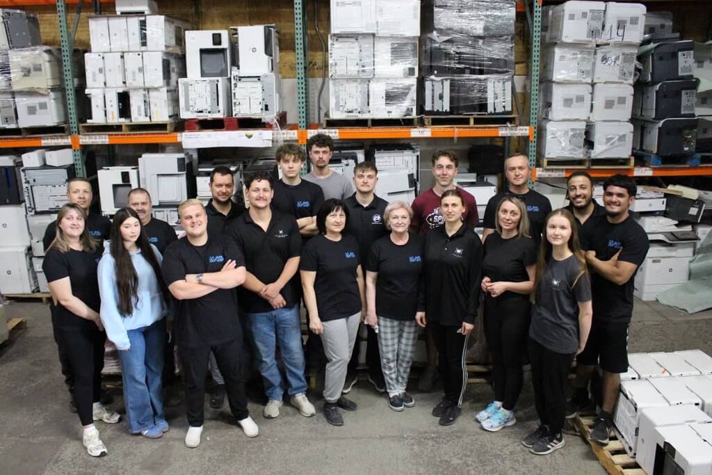 A group of 18 people, wearing mostly black shirts, stand together and smile in a warehouse filled with refurbished printers and office equipment. They are posing for a group photo.