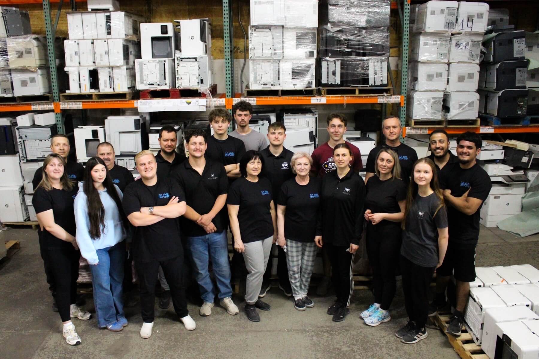 A group of people in matching logo shirts stand together in a warehouse, surrounded by shelves stacked with refurbished printers and office equipment. They are facing the camera and smiling.